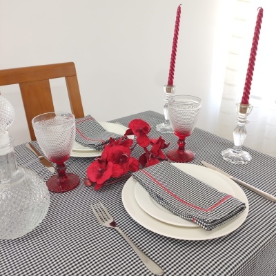 Mesa posta com toalha xadrez preta e branca, copos vermelhos, pratos brancos, guardanapos xadrez com linha vermelha, flores vermelhas e castiçais com velas vermelhas.