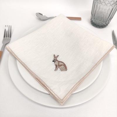 Guardanapo branco bordado com coelho sobre prato branco, talheres e copo de vidro