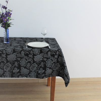 Toalha de mesa preta com padrão floral cinza numa mesa de madeira clara com prato, talheres e copo.