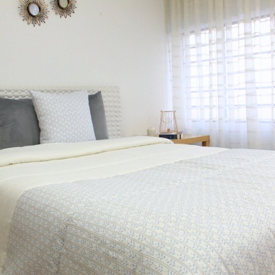 Cama com colcha padrão azul claro, almofadas cinzentas e brancas, cabeceira branca e mesa de madeira com candeeiro.
