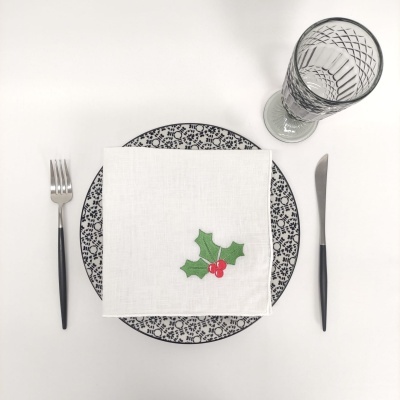 Mesa posta com prato azul decorado, guardanapo branco com detalhe de folha verde e frutos vermelhos, talheres pretos e copo transparente