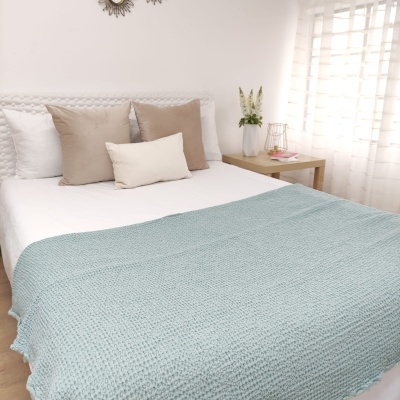 Cama com cobertor azul claro e almofadas bege e castanho claro junto a mesa de cabeceira com vaso de flores e candeeiro