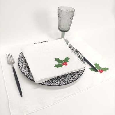 Conjunto de jantar com prato decorado, talheres pretos, taça de vidro e tecidos brancos com folhas verdes e bagos vermelhos.