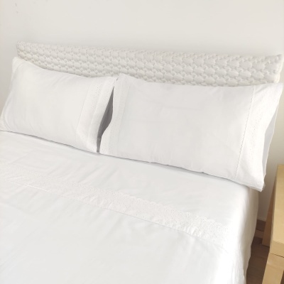 Cama com conjunto de roupa de cama branca com detalhes em renda, cabeceira texturizada branca e criado-mudo de madeira clara