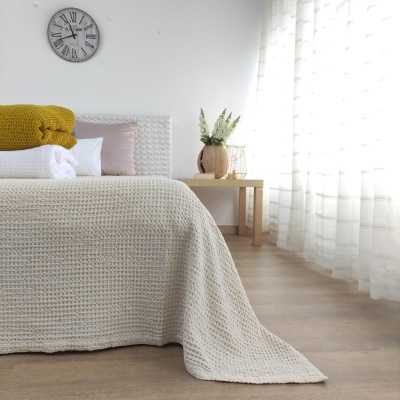 Quarto com cama e colcha creme, manta amarela, mesa de madeira clara, vaso e lâmpada, relógio de parede e cortinas brancas.