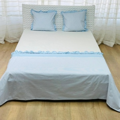 Cama de casal com roupa de cama branca e azul claro num quarto com piso em madeira e cortinas brancas