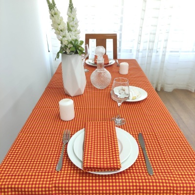 Mesa posta com toalha xadrez vermelha e amarela, pratos brancos, talheres, guardanapo, copo de vinho, vela e jarro com flores brancas