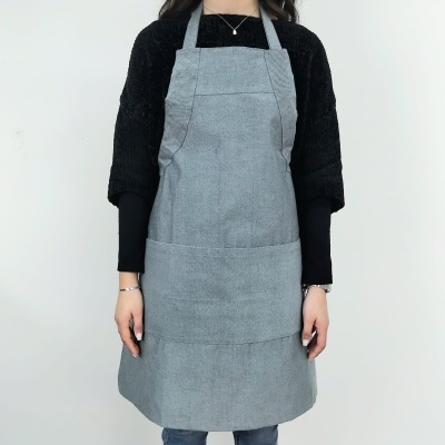 Avental cinza claro em tecido com bolso frontal dividido, usado por pessoa com camisola preta e calças azuis