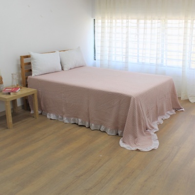 Cama com colcha rosa claro listrada e bainha branca ondulada num quarto claro com mesa de cabeceira e janela grande com cortina