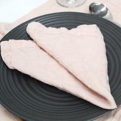 Guardanapo rosa claro dobrado sobre prato preto com colher e copo ao fundo