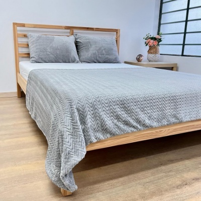 Cama de madeira clara com cobertor cinzento e almofadas e mesa de cabeceira com flores