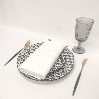 Conjunto de mesa com prato cerâmico decorado, guardanapo branco, talheres preto e dourado, e copo cinzento