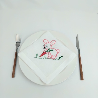 Guardanapo branco bordado com coelho e cenoura sobre prato branco, com garfo e faca de cabo de madeira