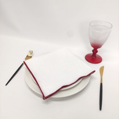 Conjunto de jantar com prato branco, guardanapo branco com borda vermelha, talheres dourados com cabos pretos e copo vermelho texturizado.