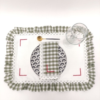 Conjunto de mesa com jogo americano branco e verde, guardanapo xadrez, prato floral, colher dourada e copo transparente.