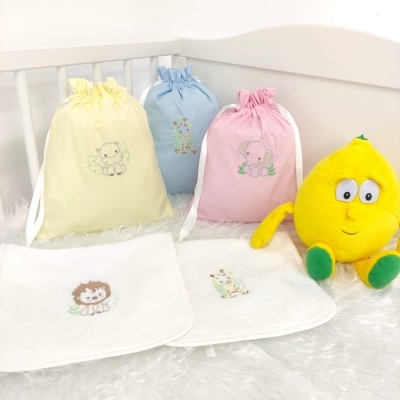 Sacos de tecido coloridos com bordados de animais, panos brancos bordados e almofada amarela em forma de limão numa cama de bebé branca