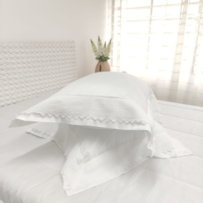 Almofada branca texturada com bordas ziguezague numa cama branca junto a vaso decorativo.