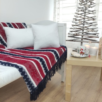 Manta decorativa azul, vermelho e branco com franjas num sofá branco junto a mesa com velas e copo decorado.