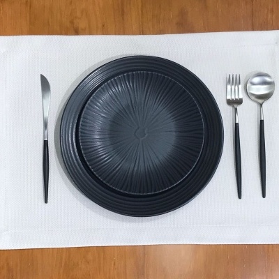 Conjunto de pratos pretos e talheres com cabo preto sobre guardanapo branco numa mesa de madeira.