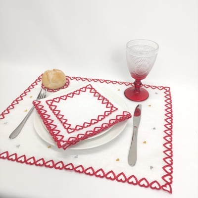 Guardanapos e toalha de mesa brancos com bordado de corações vermelhos, prato branco com pão, talheres e copo de vidro com pé vermelho num fundo branco.