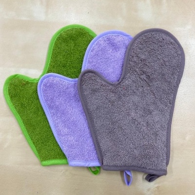 Três luvas de banho felpudas em verde, lilás e cinza sobre superfície bege