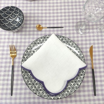 Mesa posta com prato decorado, guardanapo branco com borda roxa, talheres dourados e pretos, tigela preta com padrão branco, copos de vidro e toalha xadrez lilás.