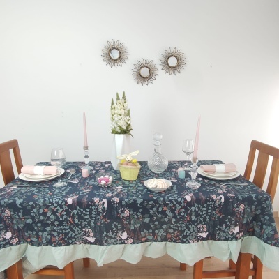 Mesa de jantar com toalha floral azul e rosa, pratos brancos, copos de vidro e decoração com flores brancas.