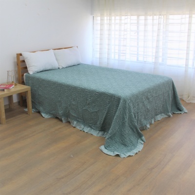 Cama com colcha verde texturada e almofadas brancas junto a mesa de cabeceira de madeira clara