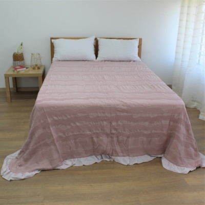 Cama de casal com edredão rosa claro e almofadas brancas numa sala com mesa de cabeceira e chão de madeira