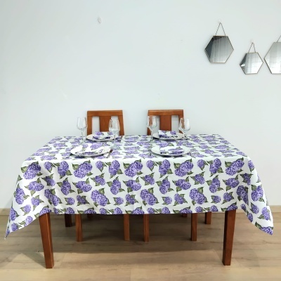 Mesa de madeira com toalha branca com padrão lilás e verde, pratos, copos e duas cadeiras de madeira