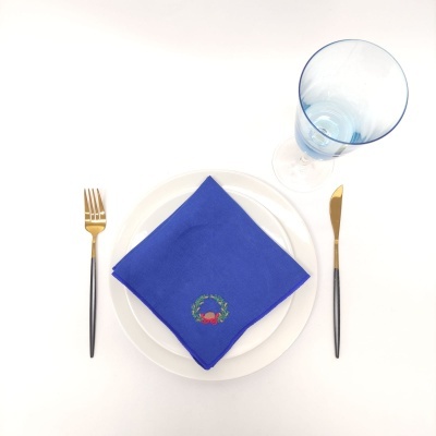 Mesa posta com talheres dourados e pretos, prato branco, guardanapo azul bordado e copo azul