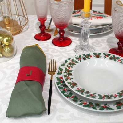 Mesa posta com conjunto de louça natalícia, talheres dourados, copos vermelhos e vela amarela.