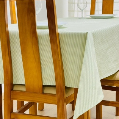 Mesa de jantar com toalha branca riscada em verde claro e cadeiras de madeira clara estofadas.