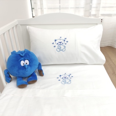 Conjunto de cama branca com ursinho bordado e peluche azul num berço branco