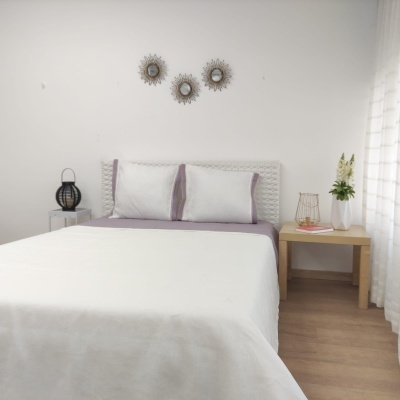 Quarto com cama de casal, roupa de cama branca e lilás, mesas de cabeceira em madeira clara e decoração com flores e espelhos