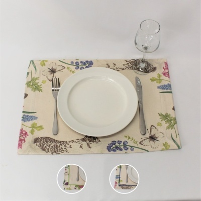 Jogo americano bege estampado com flores, folhas, borboletas e esquilo, com prato branco, talheres e taça de vidro numa mesa branca