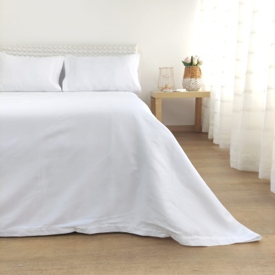 Cama com lençóis brancos e cabeceira trançada, mesa de madeira clara com jarro de flores e luminária, cortinas brancas