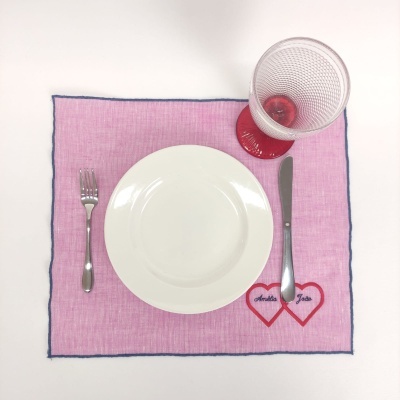 Toalhete rosa com corações vermelhos e nomes, prato branco, talheres metálicos e copo de vidro com base vermelha