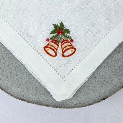 Guardanapo branco com bordado de sinetas douradas, folhas verdes e bagas vermelhas sobre prato cinzento