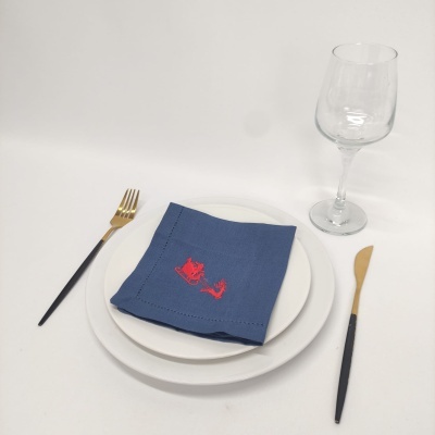 Conjunto de mesa com prato branco, guardanapo azul bordado, talheres dourados e copo de vidro