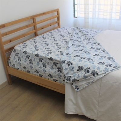 Cama de madeira clara com cabeceira e roupa de cama branca e manta estampada com folhas azuis.