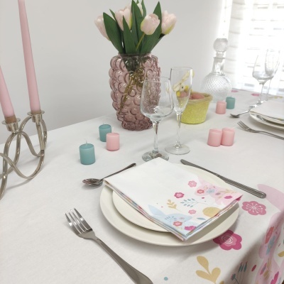 Mesa posta com toalha branca estampada, guardanapo florido, pratos, talheres, copos, vaso rosa com tulipas e velas coloridas.