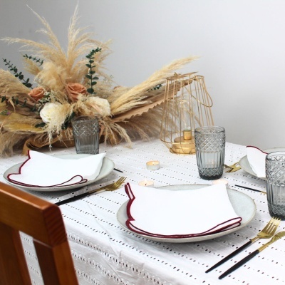 Mesa posta elegante com pratos brancos e bordas vermelhas, copos cinza, talheres dourados e arranjo floral seco.