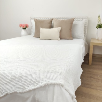 Cama com colcha branca tricotada e almofadas bege e branca, mesa de cabeceira com vaso e flores