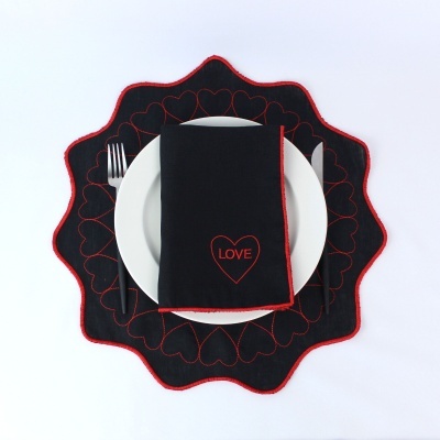 Conjunto de mesa com prato branco, guardanapo preto com LOVE vermelho e descanso de prato preto com corações vermelhos bordados