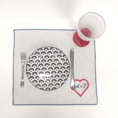 Jogo americano quadrado bege com coração bordado, prato branco com padrão preto, talheres de metal e copo transparente com base vermelha