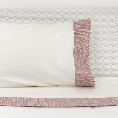 Almofada branca com borda rosa sobre cama com lençol e cabeceira texturizados