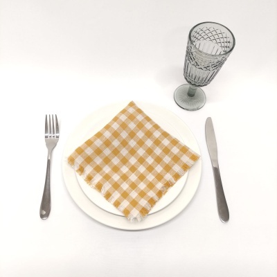 Mesa posta com pratos brancos, guardanapo amarelo xadrez, talheres metálicos e copo cinzento translúcido