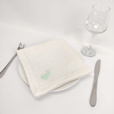 Guardanapo branco com bordado verde sobre prato branco, garfo, faca e taça de vidro