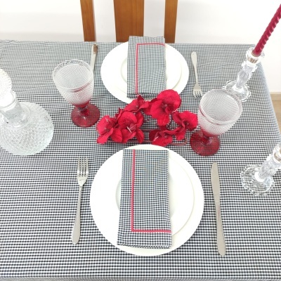 Mesa posta com pratos, guardanapos xadrez, talheres, taças vermelhas, castiçais de vidro e flores vermelhas